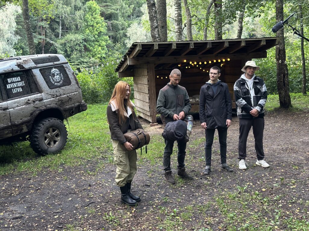 viena sieviete un trīs vīrieši stāv mežā, aiz viņiem ir koks un kravas auto