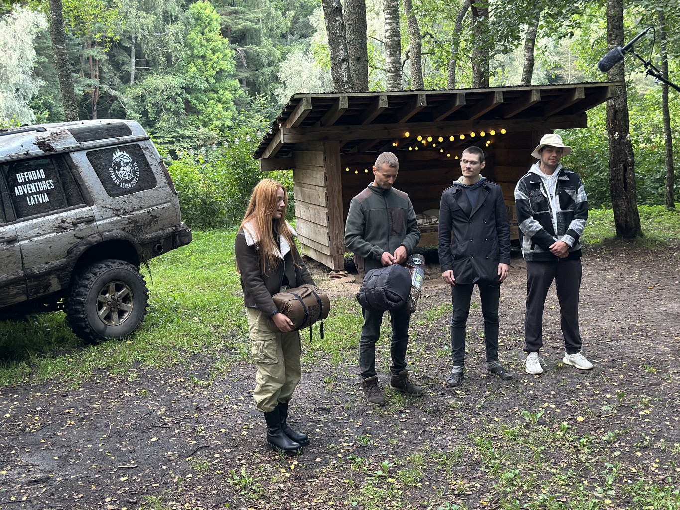 viena sieviete un trīs vīrieši stāv mežā, aiz viņiem ir koks un kravas auto