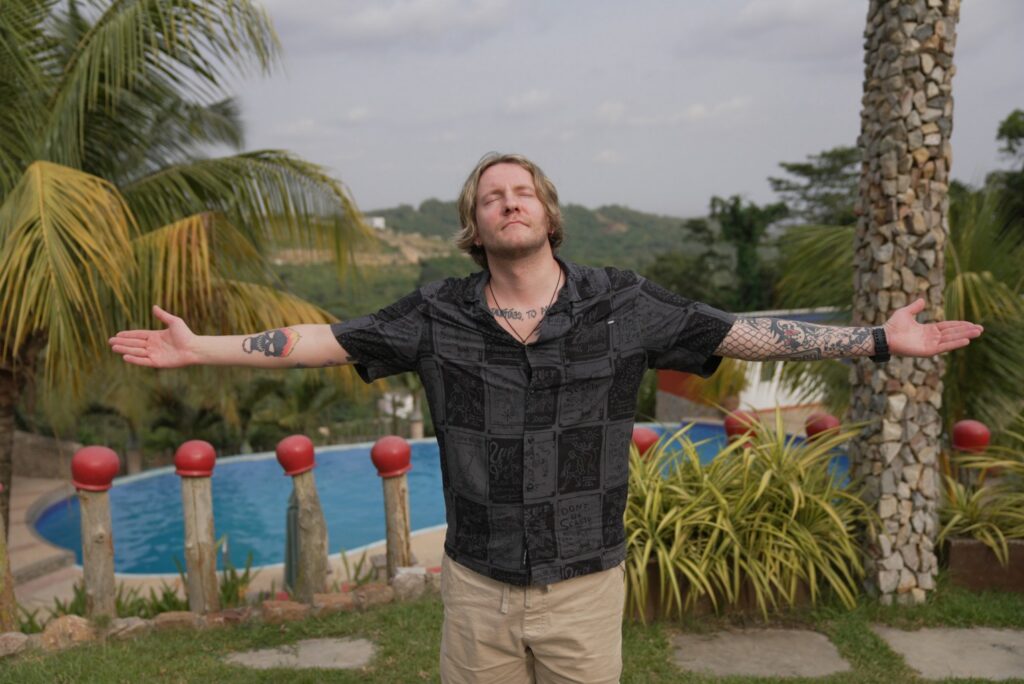 Vīrietis raugās augšup debesīs ar rokām sānos, aiz viņa redzama Āfrikas ainava