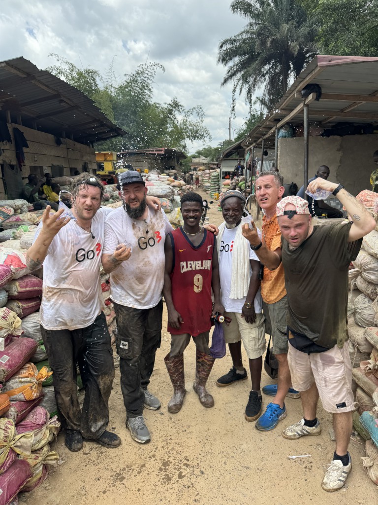 Cilvēki stāv ārā pie tirgus, daži no tiem valkā GO3 T-kreklus