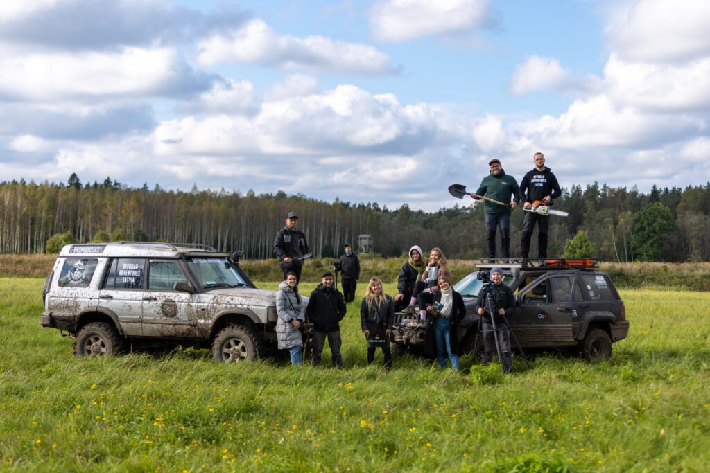 daudzi cilvēki mežā blakus lielām automašīnām skatās uz kameru
