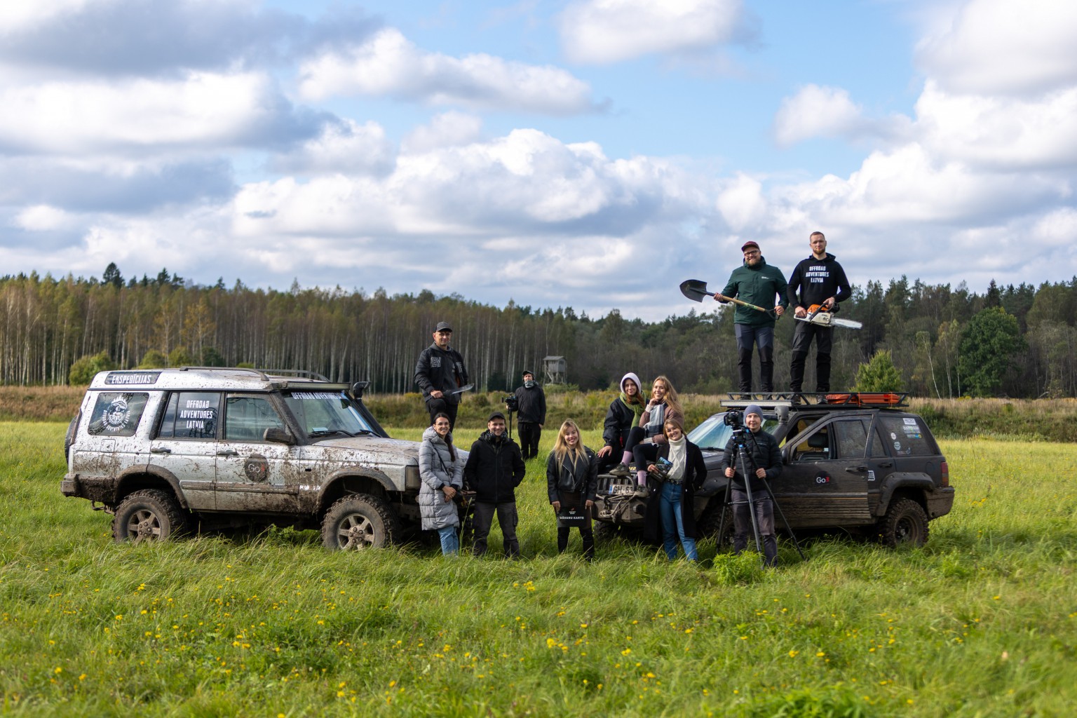 Cilvēku grupa stāv uz zāles pie meža un divām automašīnām