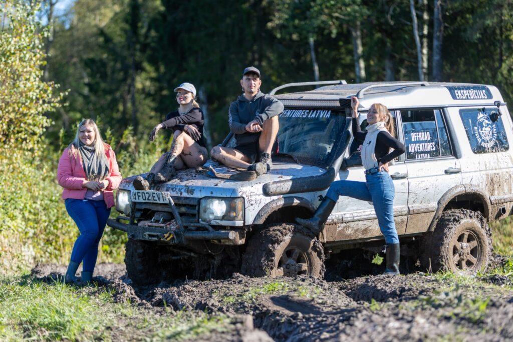 Trīs sievietes un viens vīrietis sēž vai atspiežas pie kravas automašīnas uz dubļainas zāles