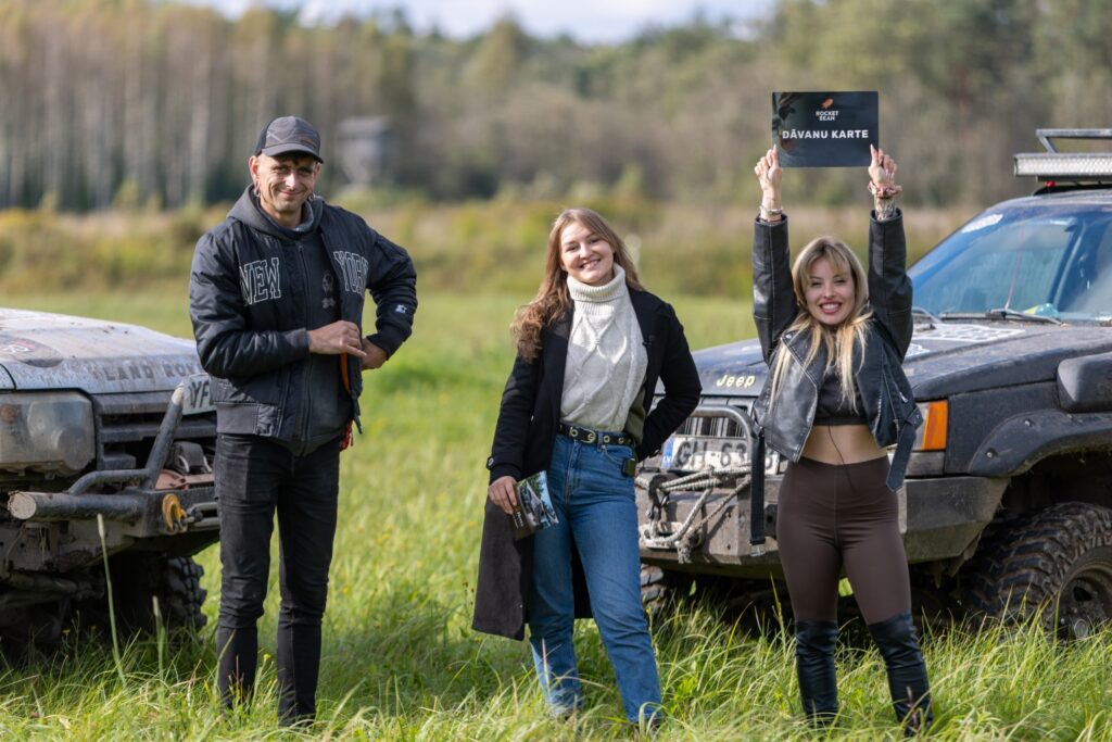 Divas sievietes un viens vīrietis stāv pie kravas automašīnām uz zāles