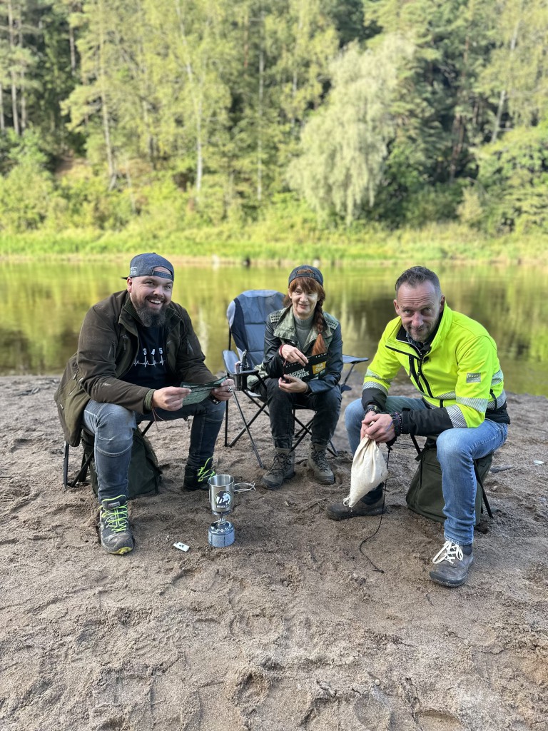 Divi vīrieši un viena sieviete sēž krēslos pie upes