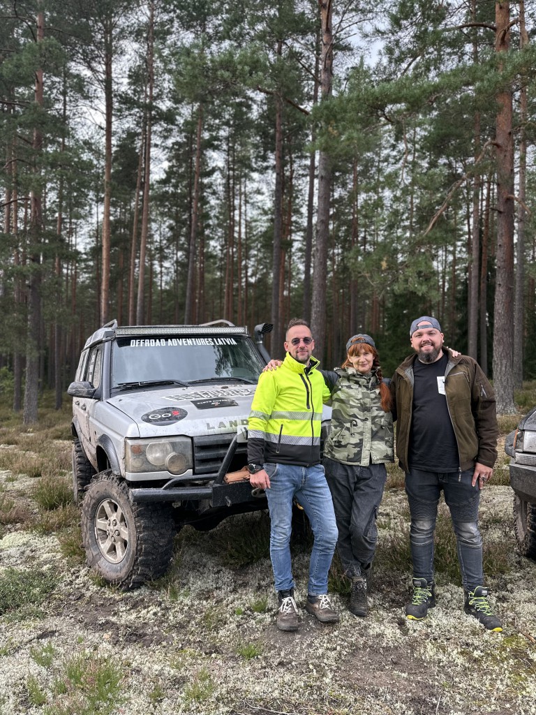 Viena sieviete un divi vīrieši stāv pie kravas automašīnas un mežā