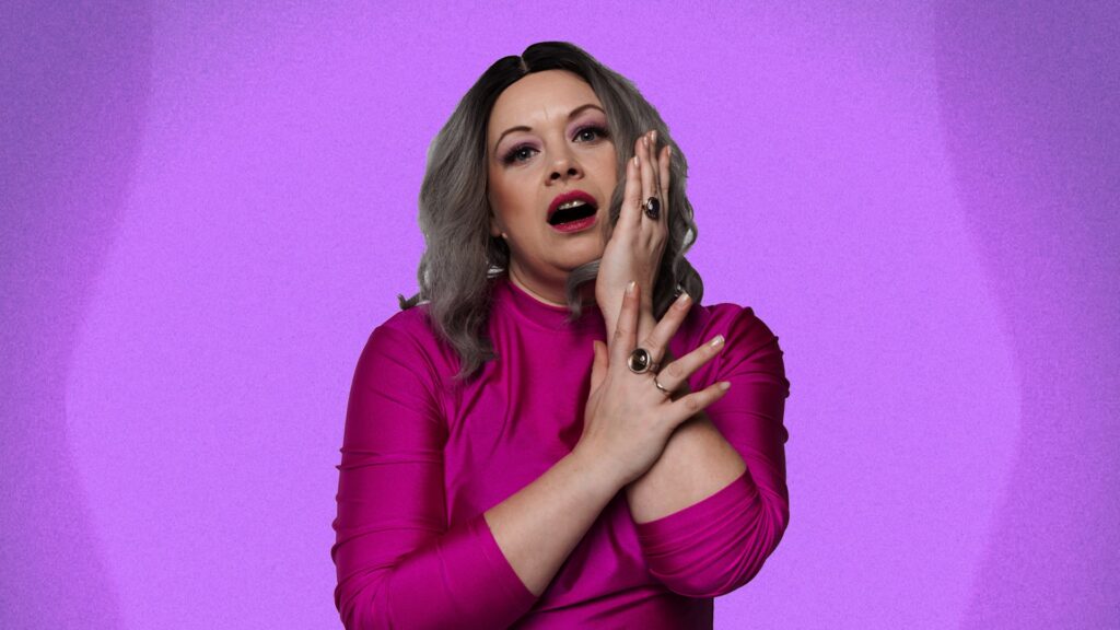 Blonda sieviete rozā apģērbā uz violetas fona, skatās tieši priekšā.